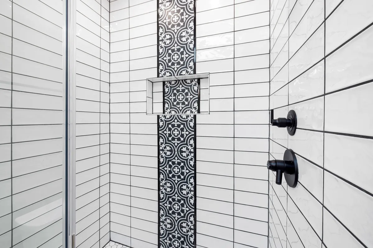 A shower wall with white tiles, dark grout and a patterned decorative strip