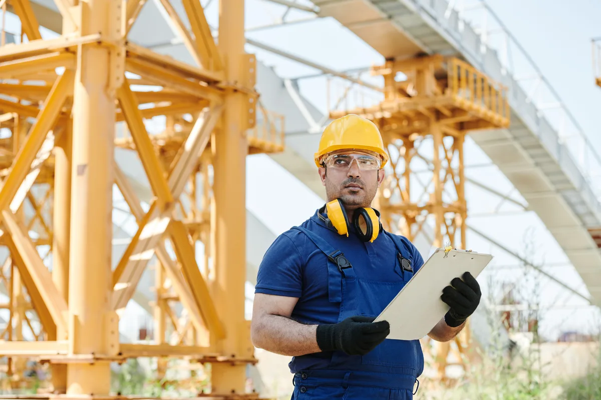 Tradesman with a yellow helmet, safety glasses, earmuffs around his neck and black work gloves holding a clipboard