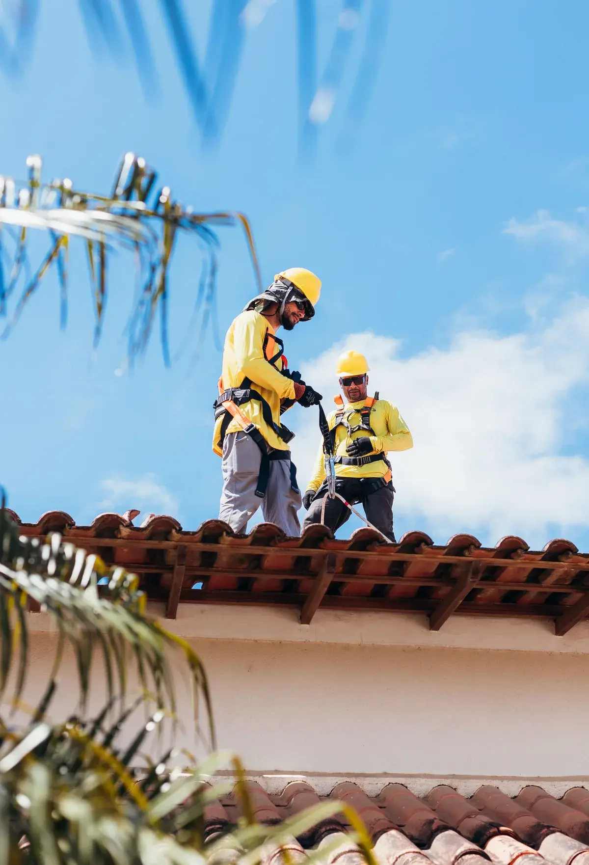 Two roofers wearing yellow helmets, gloves and fall-arrest harnesses at work on a tiled roof