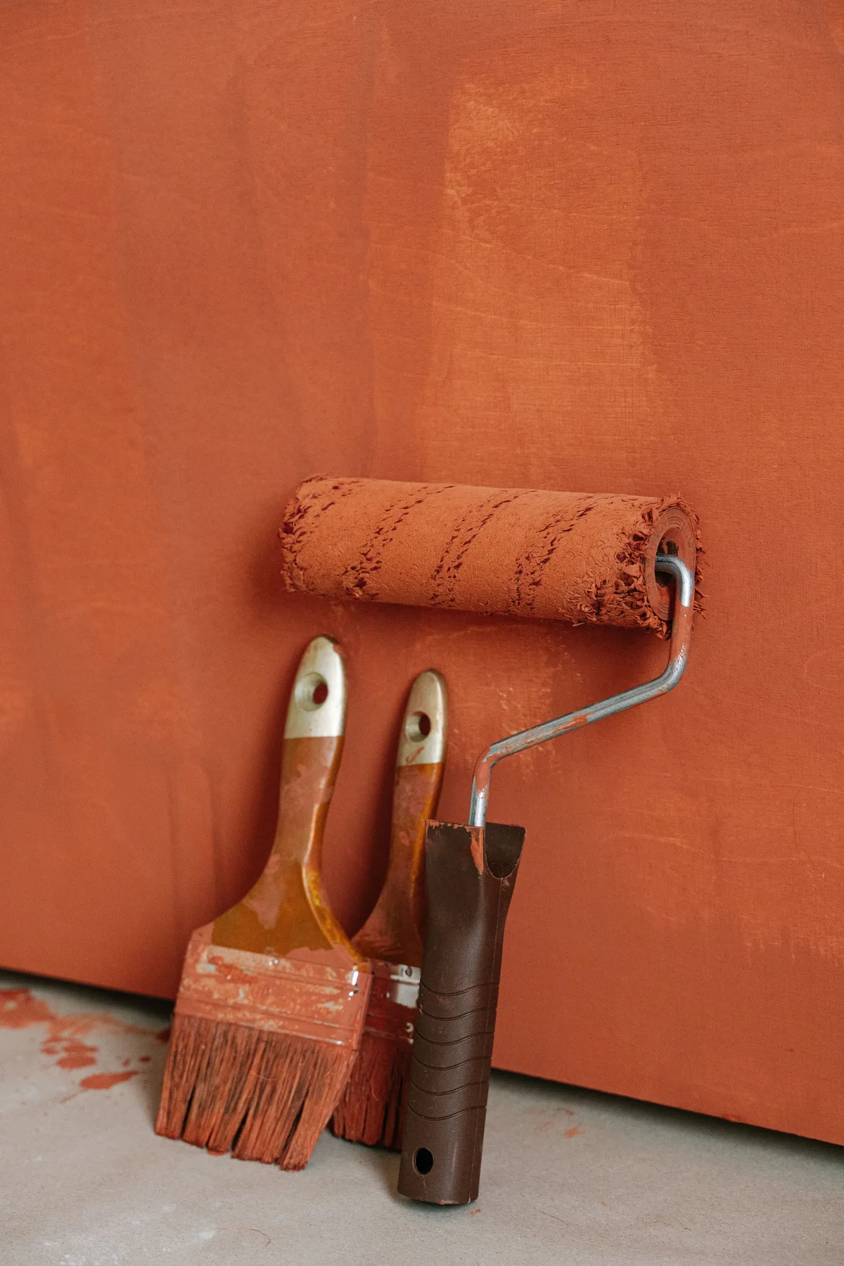 Gebruikte verfroller en twee kwasten met oranje verfresten tegen een geschilderde muur