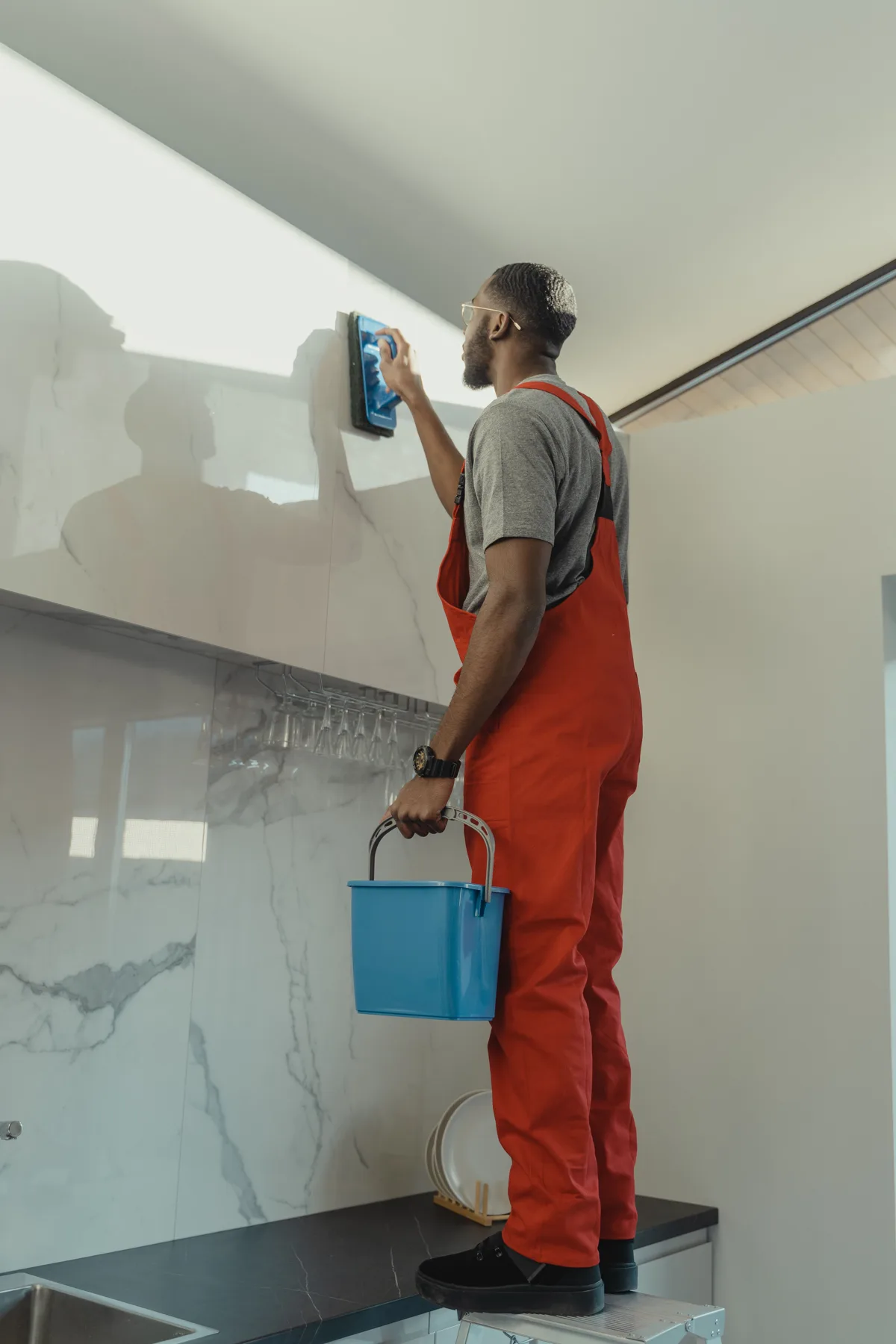 A man in red overalls cleans a white marble-look wall tile with a blue sponge