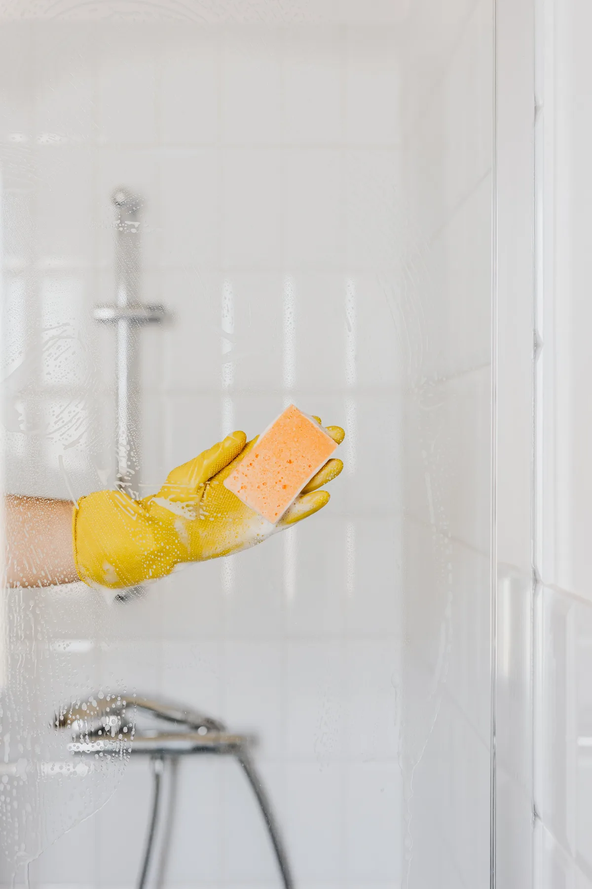 Hand in a yellow glove scrubbing a soapy glass shower screen with a sponge