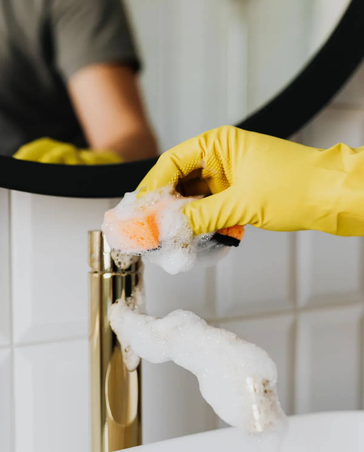 Hand in gele huishoudhandschoen poetst een goudkleurige kraan met een spons en schuim