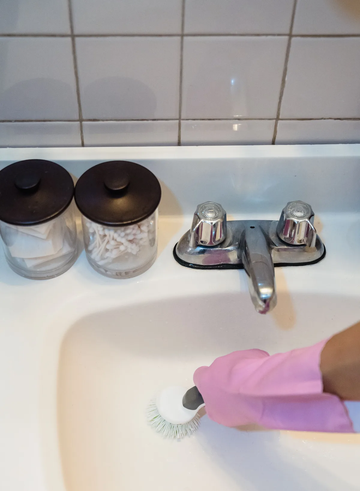 Ouderwetse wastafelkraan met twee knoppen boven een witte wastafel, een hand met roze handschoen borstelt de wastafel schoon