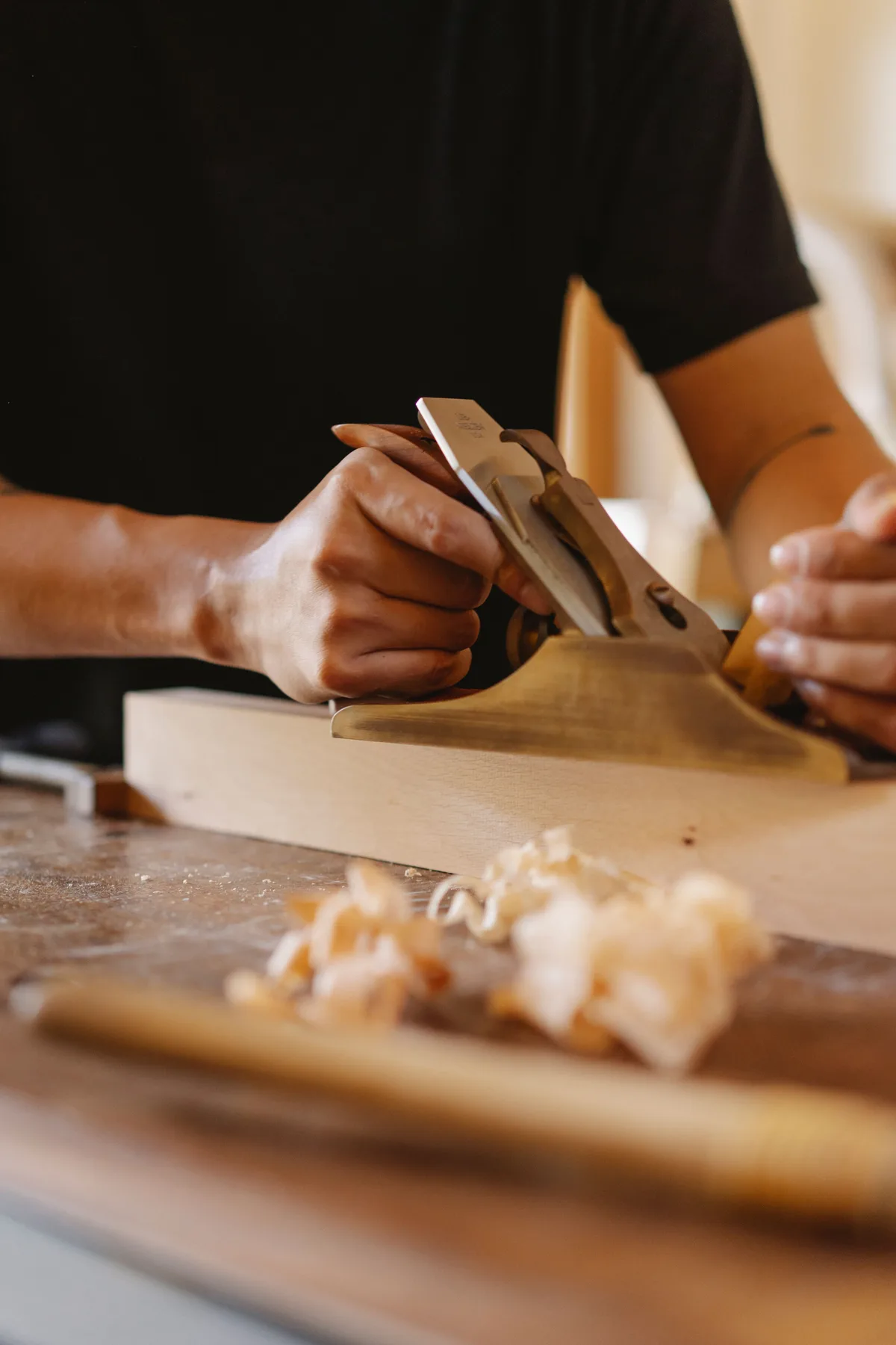 Two hands planing a wooden board with a hand plane, with shavings on the workbench