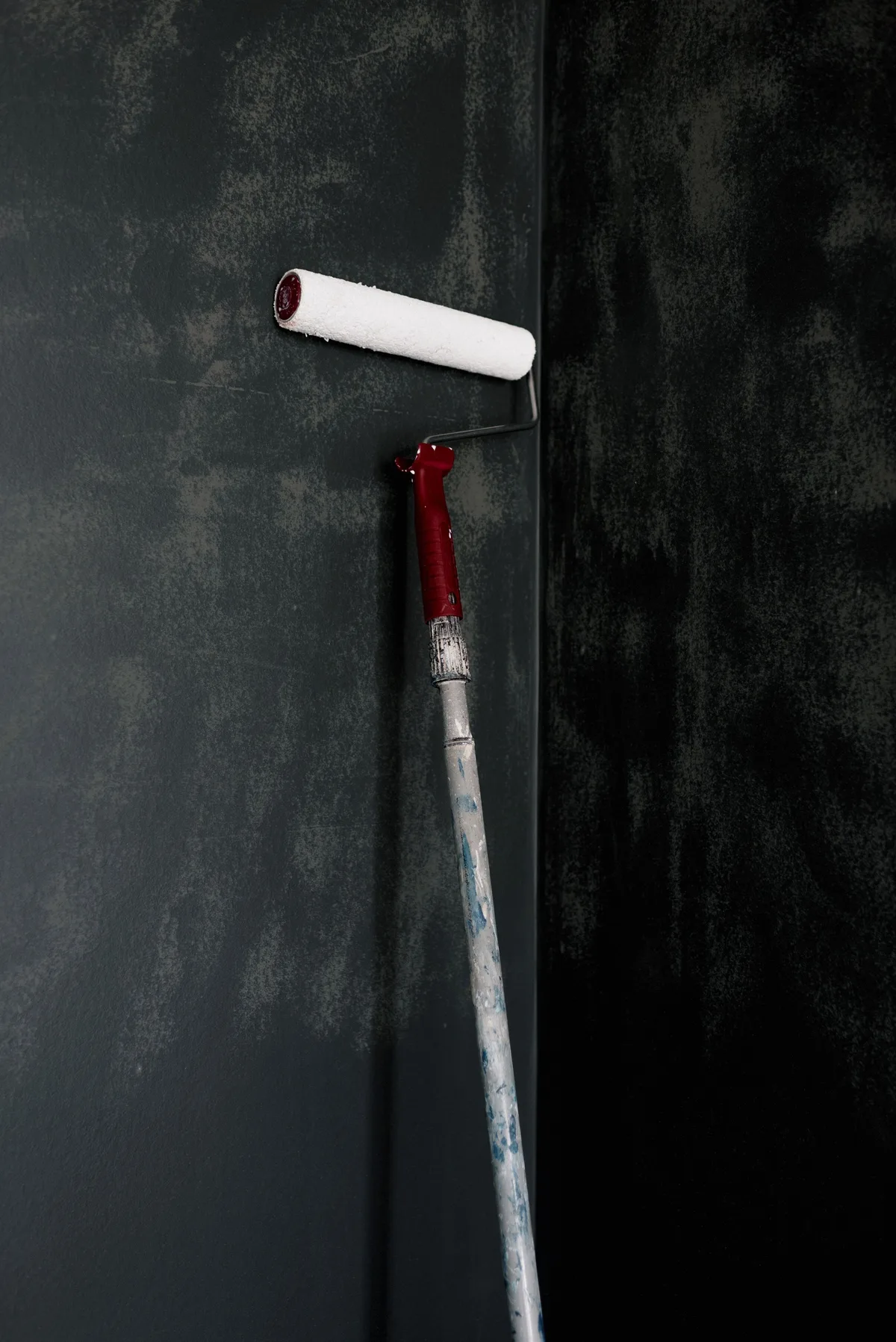 A paint roller with a white sleeve on a red pole leans against a dark wall