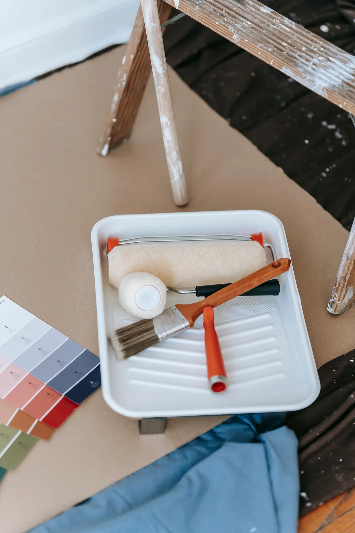 A paint tray with a roller and a brush on masking paper, next to the legs of a painter's ladder