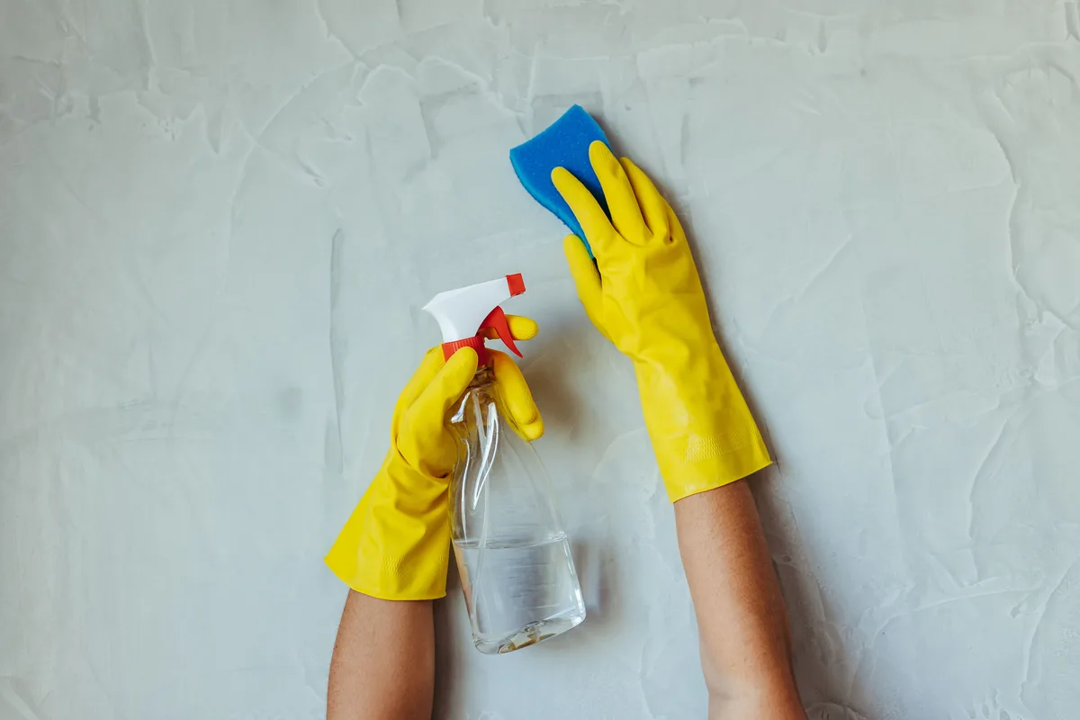 Hands in yellow household gloves cleaning a wall with a spray bottle and a blue sponge