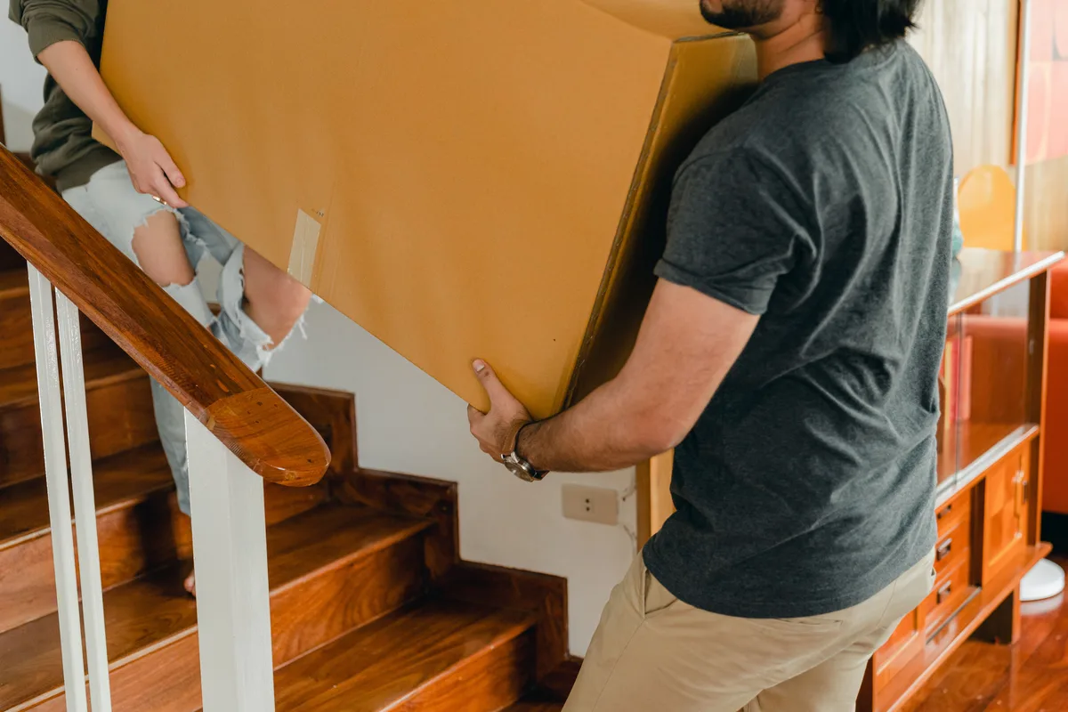 Two people carrying a large cardboard box together along a staircase in a home