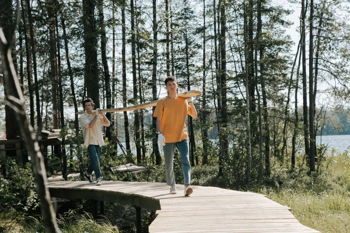 Two people carrying a long wooden plank along a boardwalk through a forest by a lake