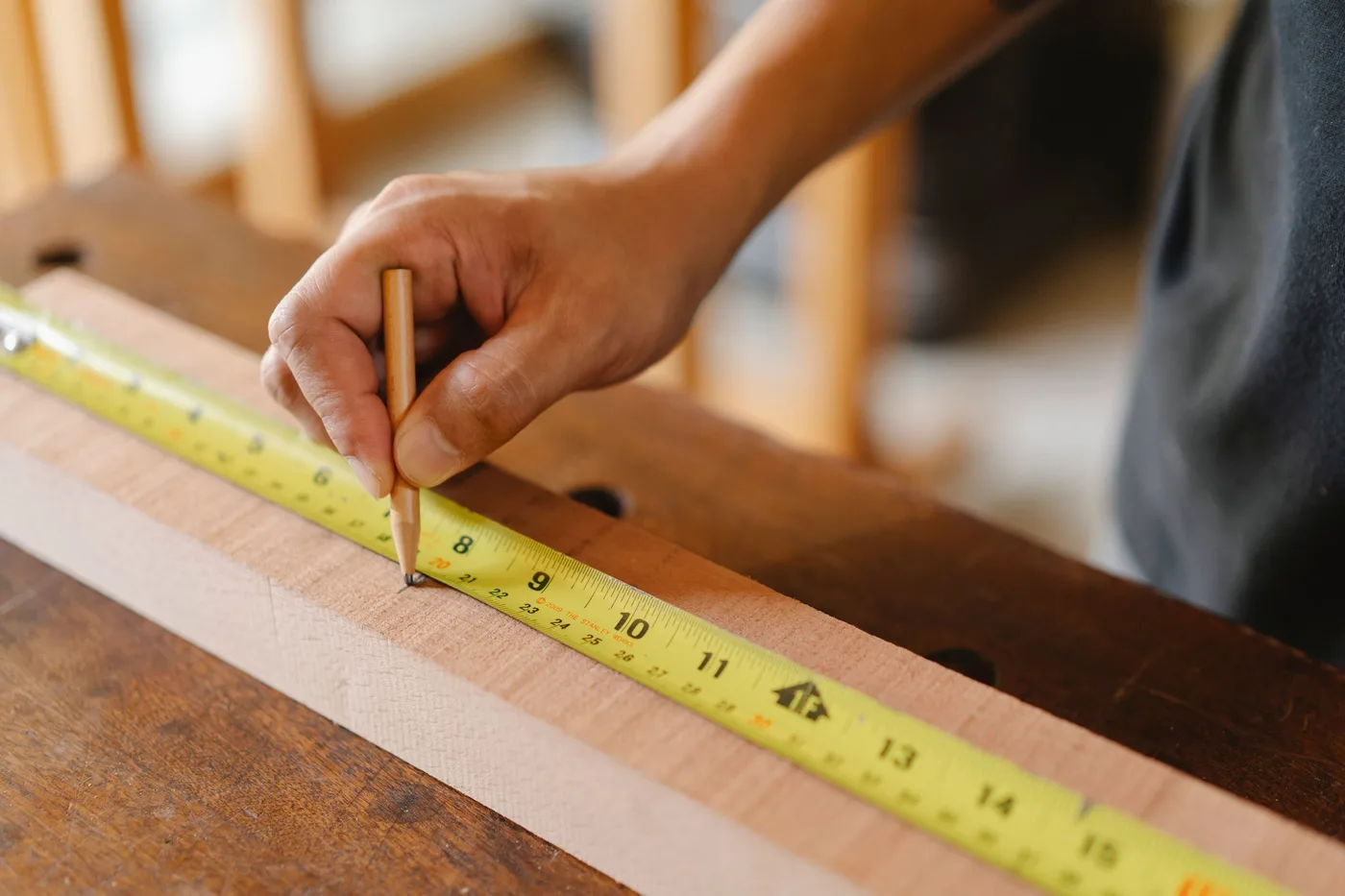 A hand marking a measurement with a pencil along a yellow tape measure on a wooden board