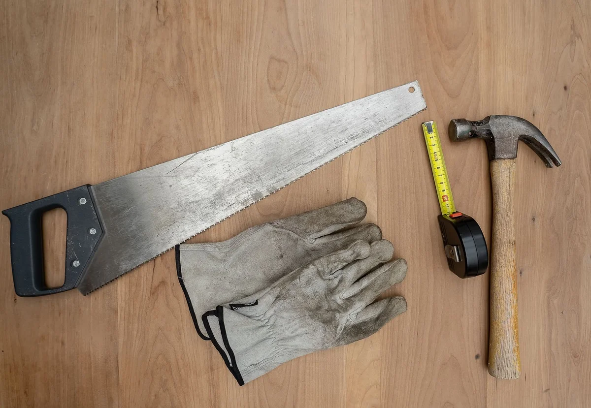 A hand saw, work gloves, a tape measure and a hammer on a wooden surface