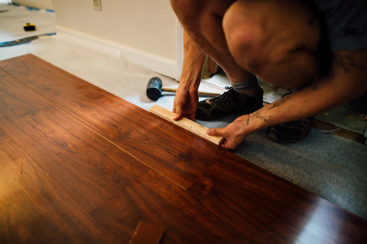 A DIYer laying laminate on a grey underlay with a wooden spacer batten and a rubber hammer