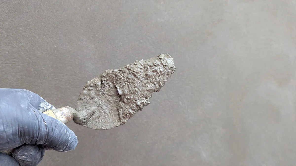 A gloved hand holds a trowel with plaster against a smooth grey wall