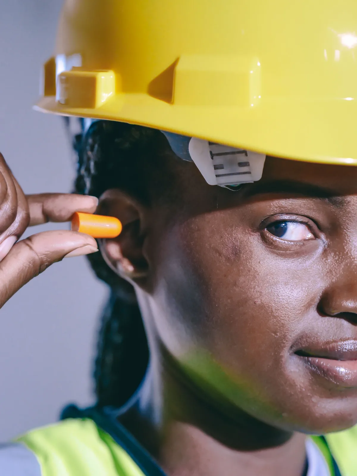 Vrouw met gele veiligheidshelm en geel hesje houdt een oranje schuimoordopje bij haar oor