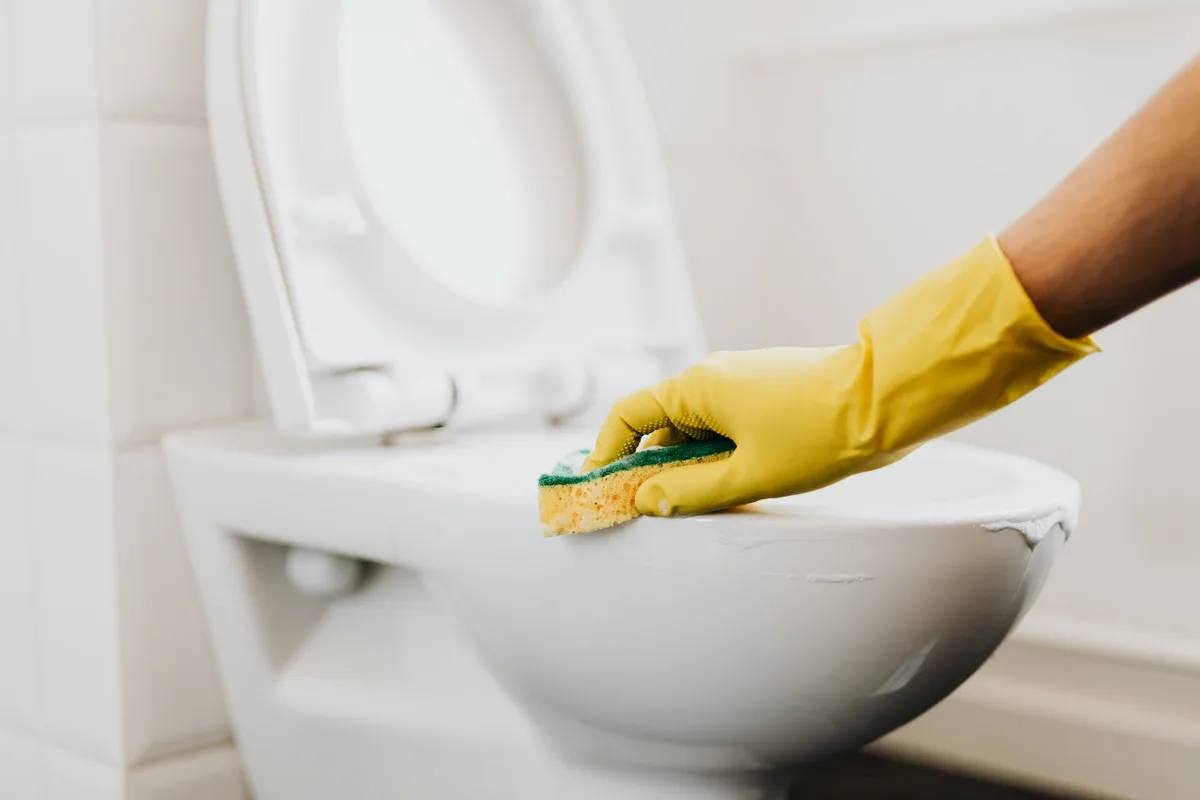 Hand in a yellow household glove cleaning the rim of a white toilet with a sponge