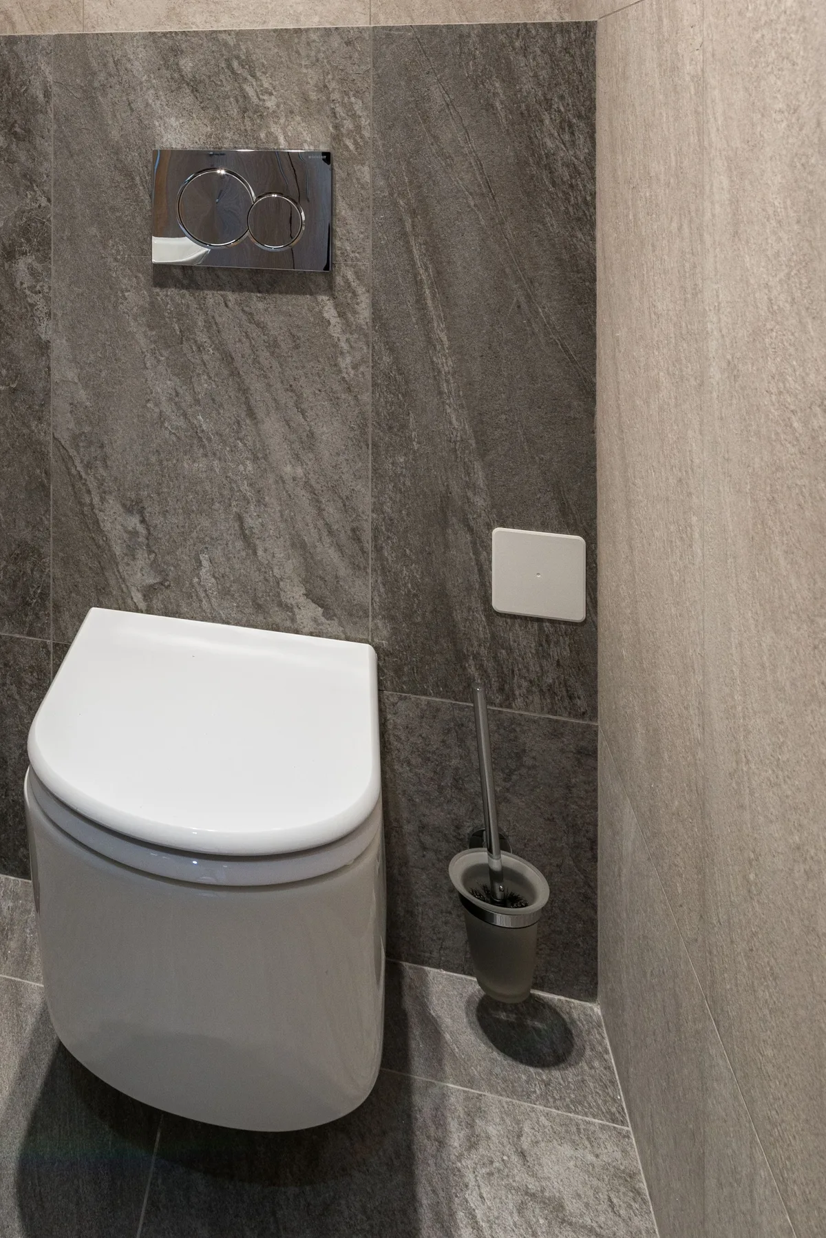 Toiletruimte met wit hangtoilet en chromen bedieningsplaat op een donkere tegelwand