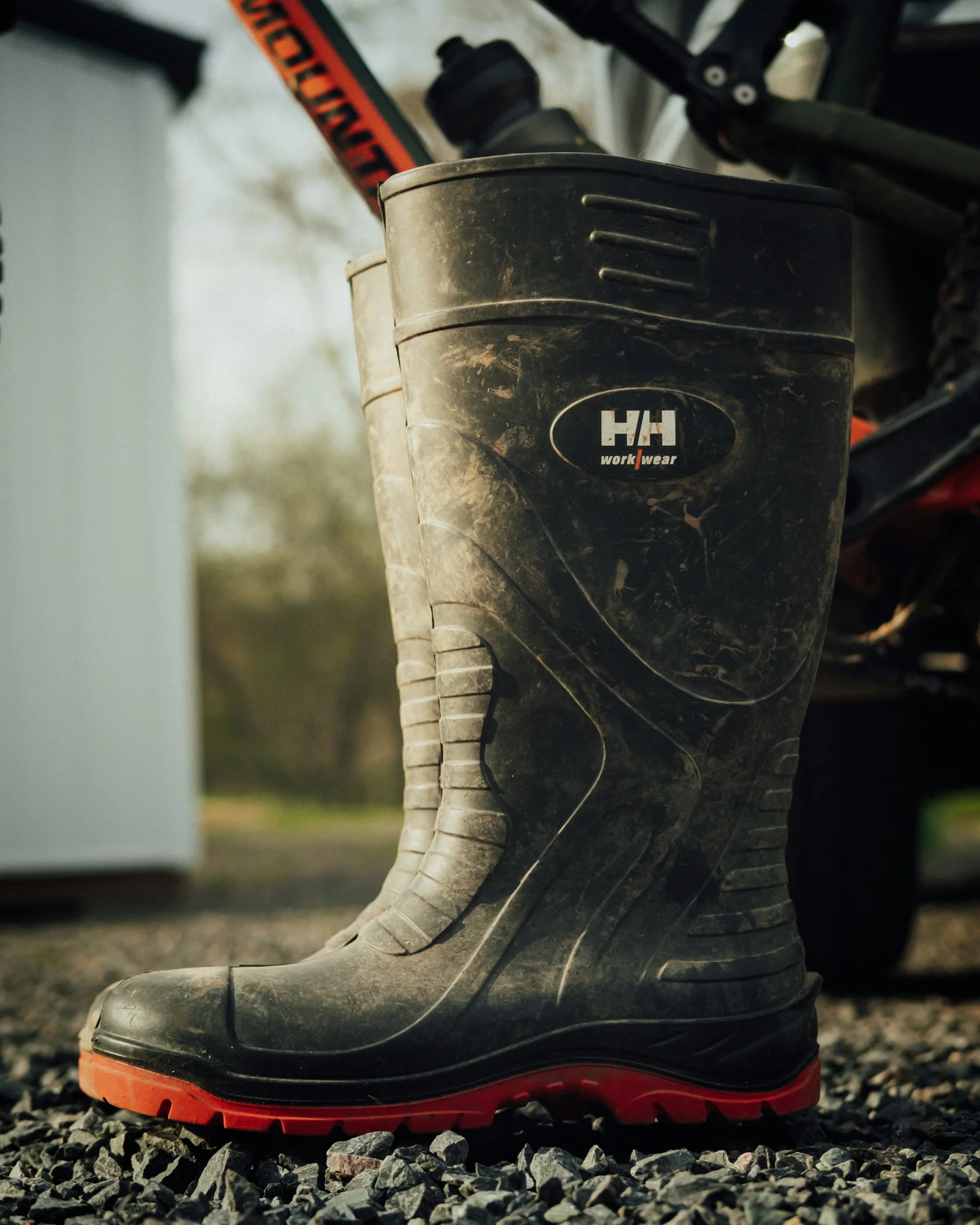 Muddy black rubber work boot with a red sole on gravel