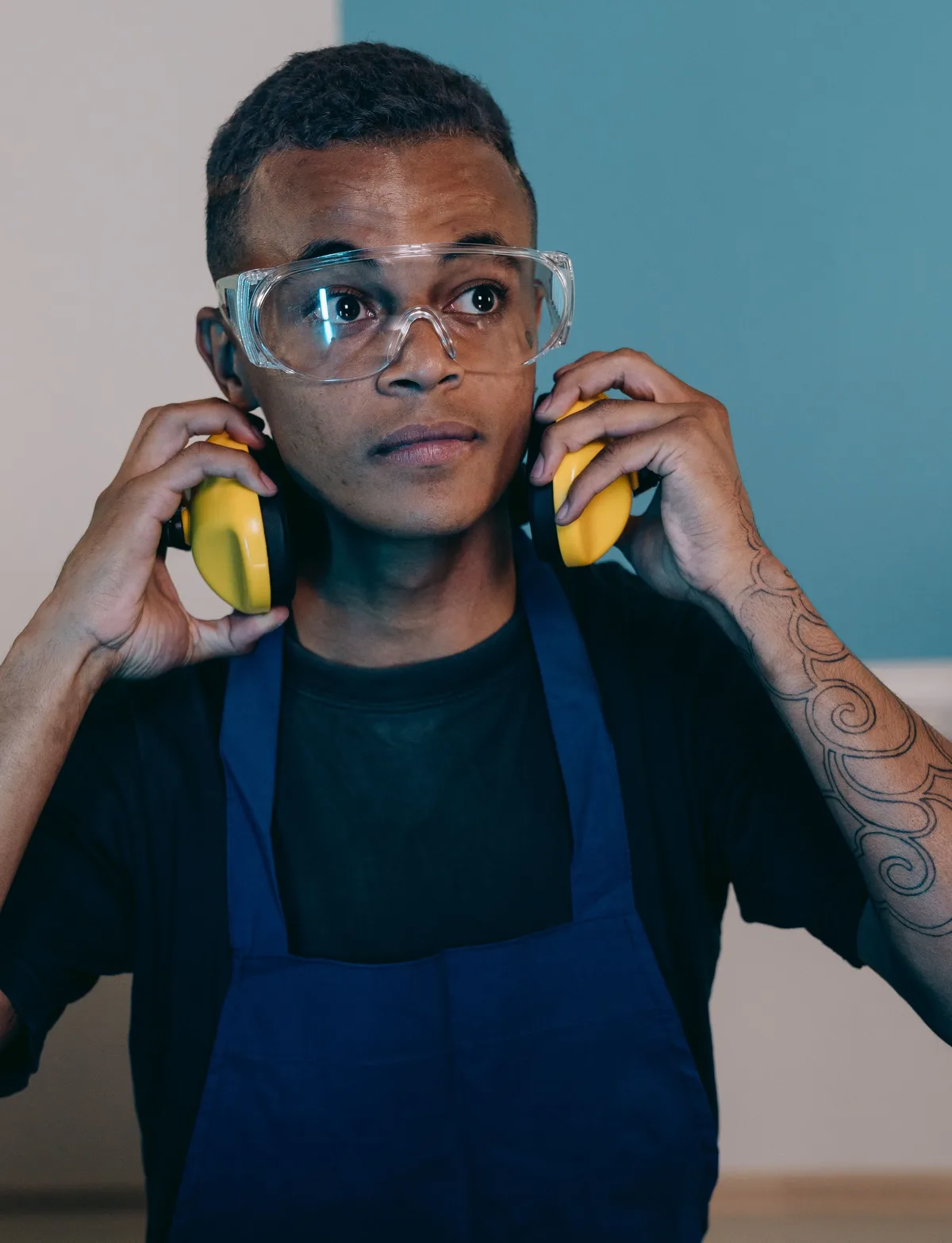 Man wearing clear wide-vision safety glasses puts on yellow earmuffs in a workshop
