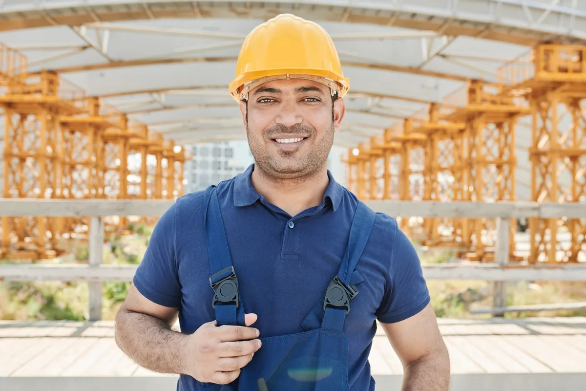 Professional wearing a yellow safety helmet and blue bib overalls on a site with scaffolding