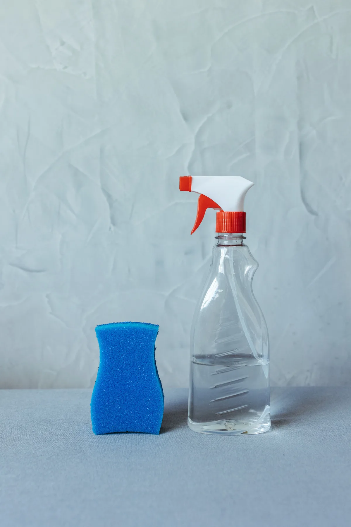 A blue sponge and a transparent spray bottle of water side by side against a light grey wall