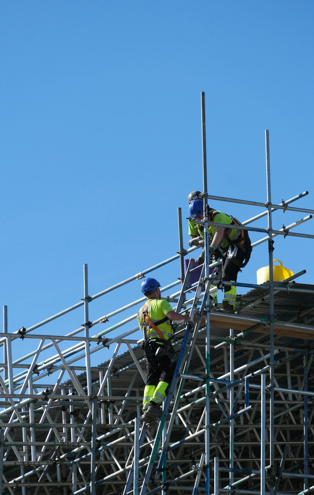 Two construction workers wearing helmets, high-visibility clothing and safety harnesses climbing scaffolding