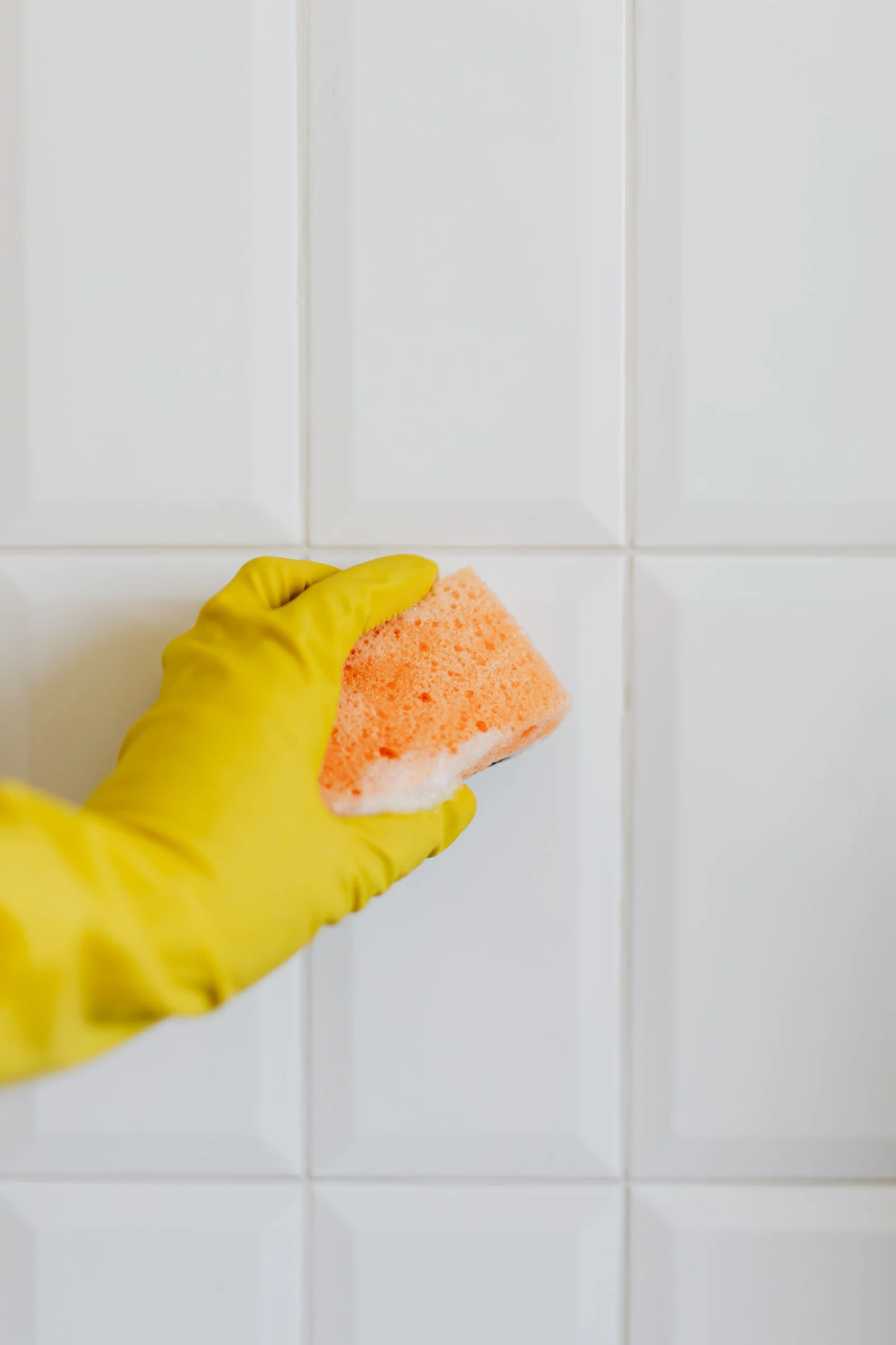 A hand in a yellow rubber glove cleans a white wall tile with an orange sponge