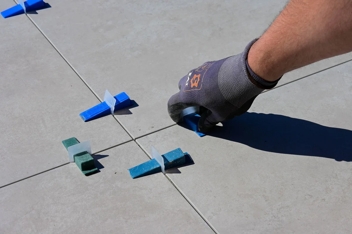 A gloved hand places a levelling clip between large floor tiles