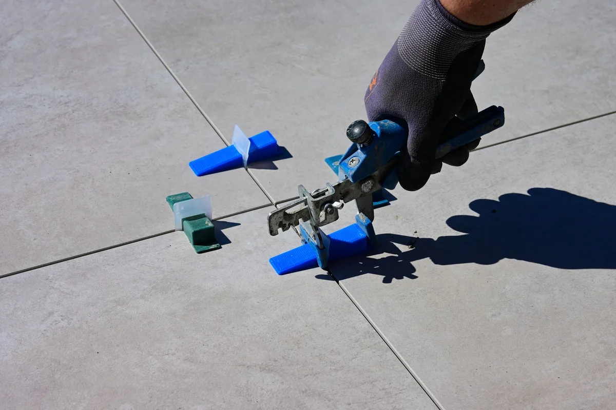A gloved hand uses a tile levelling tool on large grey floor tiles