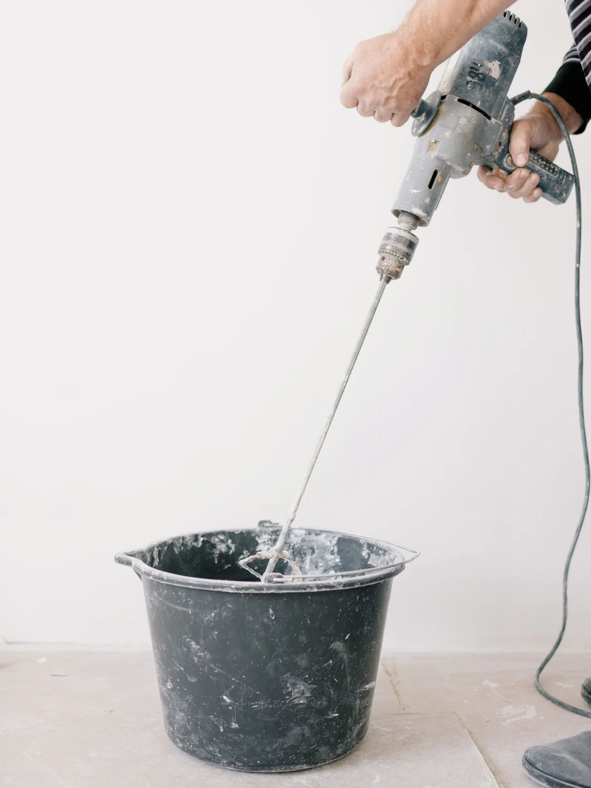 A DIYer mixing tile adhesive in a bucket with a drill and mixing paddle