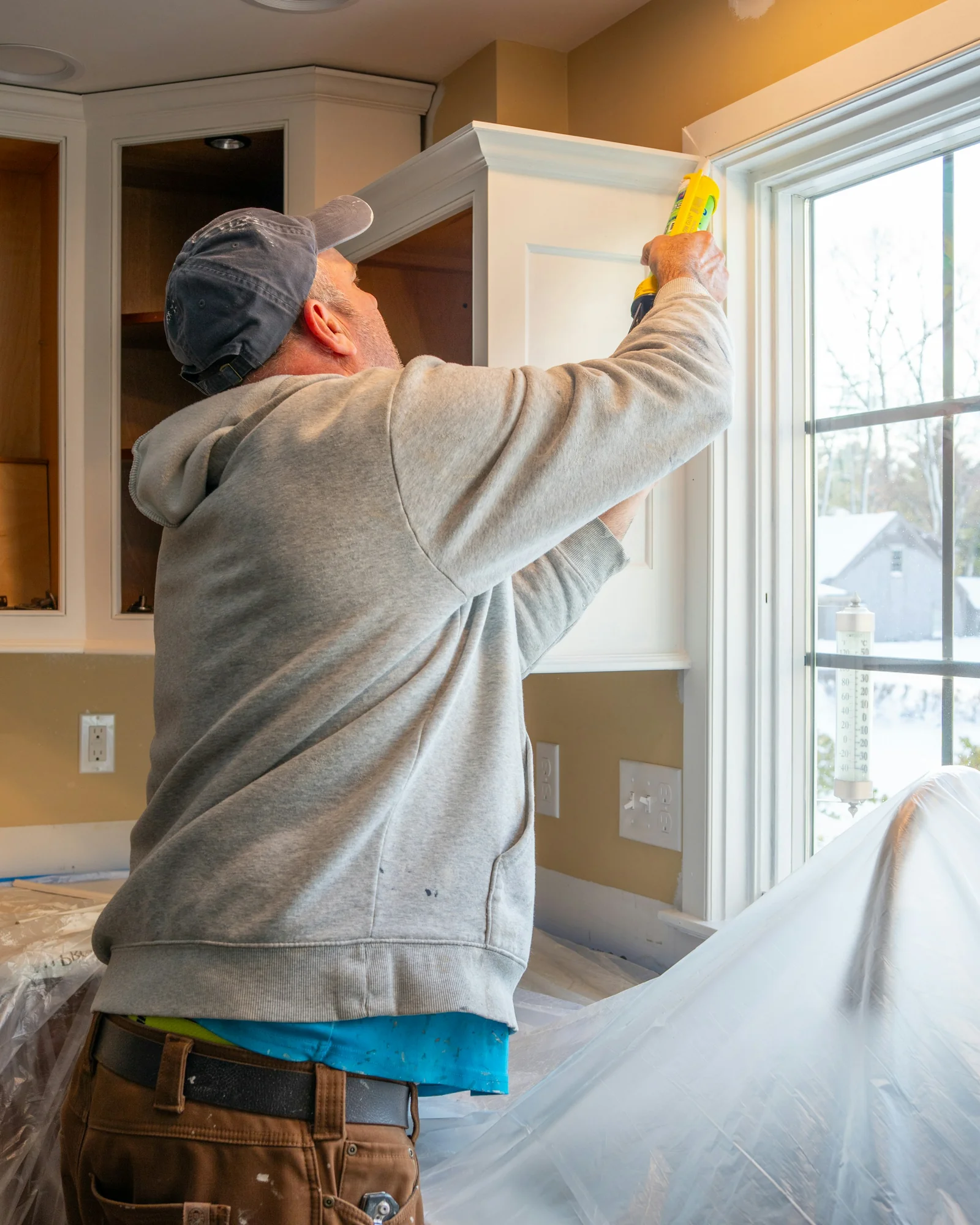 A handyman applying sealant along a white window frame in a home