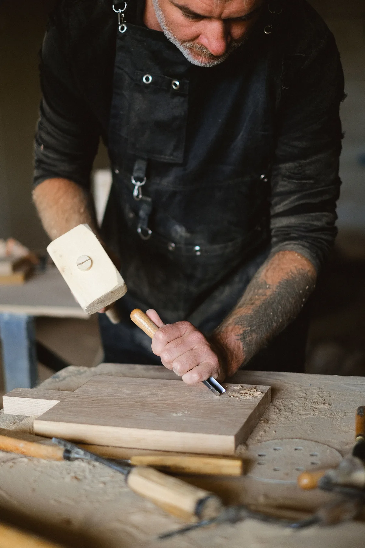 Woodworker in a work apron cutting a recess in a board on the workbench with a wood chisel and a wooden mallet, with more chisels in the foreground