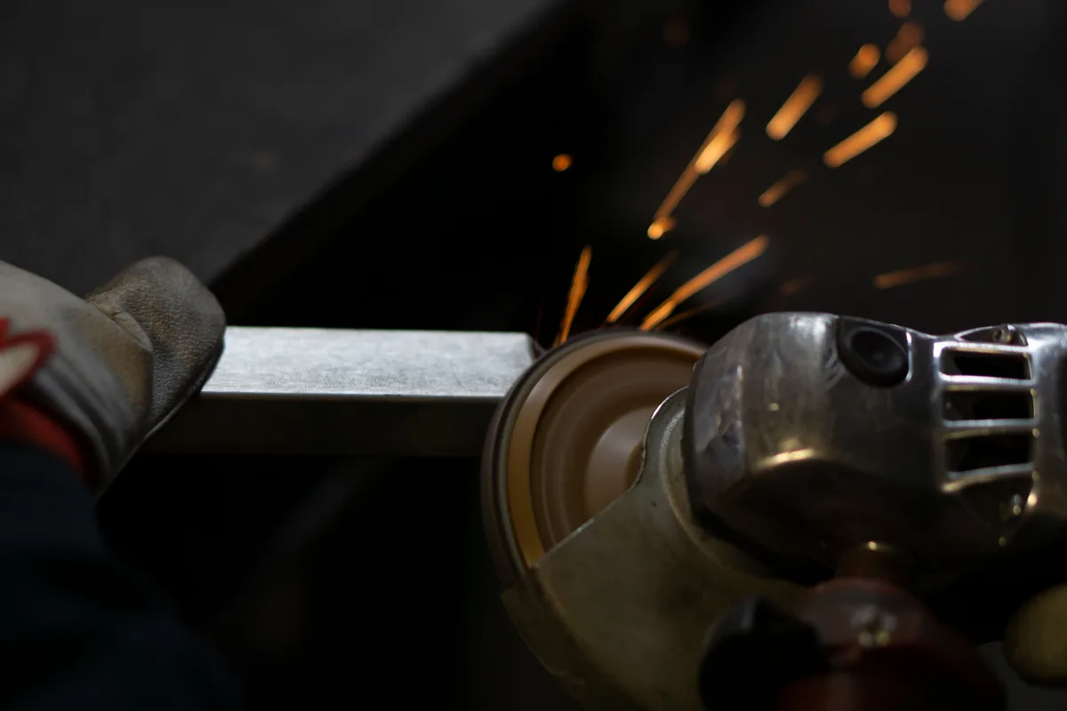 Close-up of an angle grinder with a guard grinding a steel box section, a gloved hand steadying the section away from the disc and sparks flying